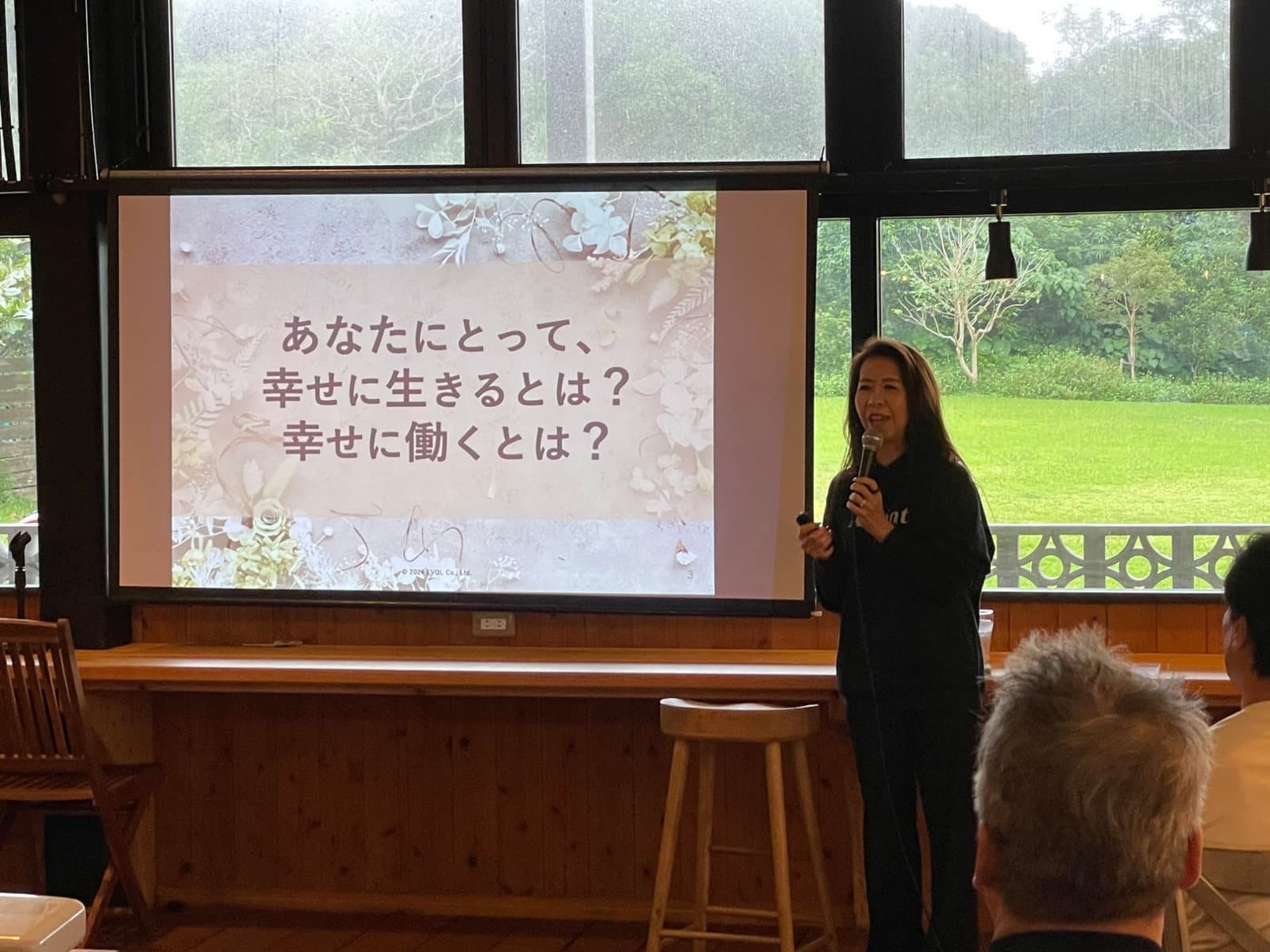 沖縄ウェルビーイング・リトリートで登壇する前野マドカさん。スクリーンには「あなたにとって、幸せに生きるとは？ 幸せに働くとは？」と映っている
