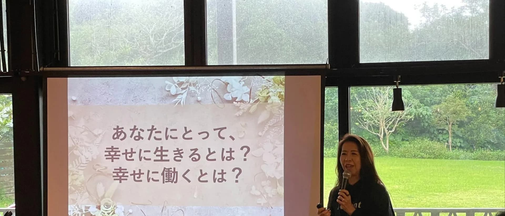 研修会場のスクリーンに「あなたにとって、幸せに生きるとは？ 幸せに働くとは？」と映し出され、その前で話す人がいる。窓の外には緑が広がっている