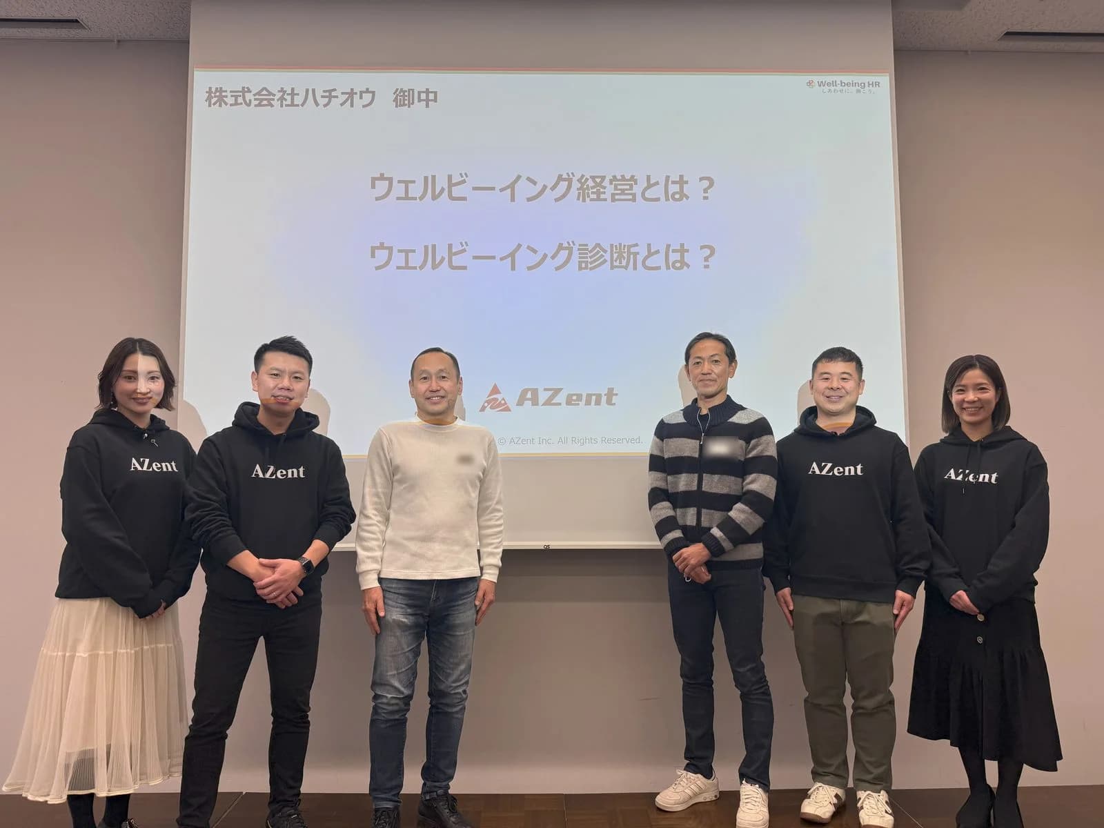 「株式会社ハチオウ 御中 ウェルビーイング経営とは? ウェルビーイング診断とは?」と映し出されたスクリーンの前に、AZentのメンバーとハチオウのご担当者が並んで立っている